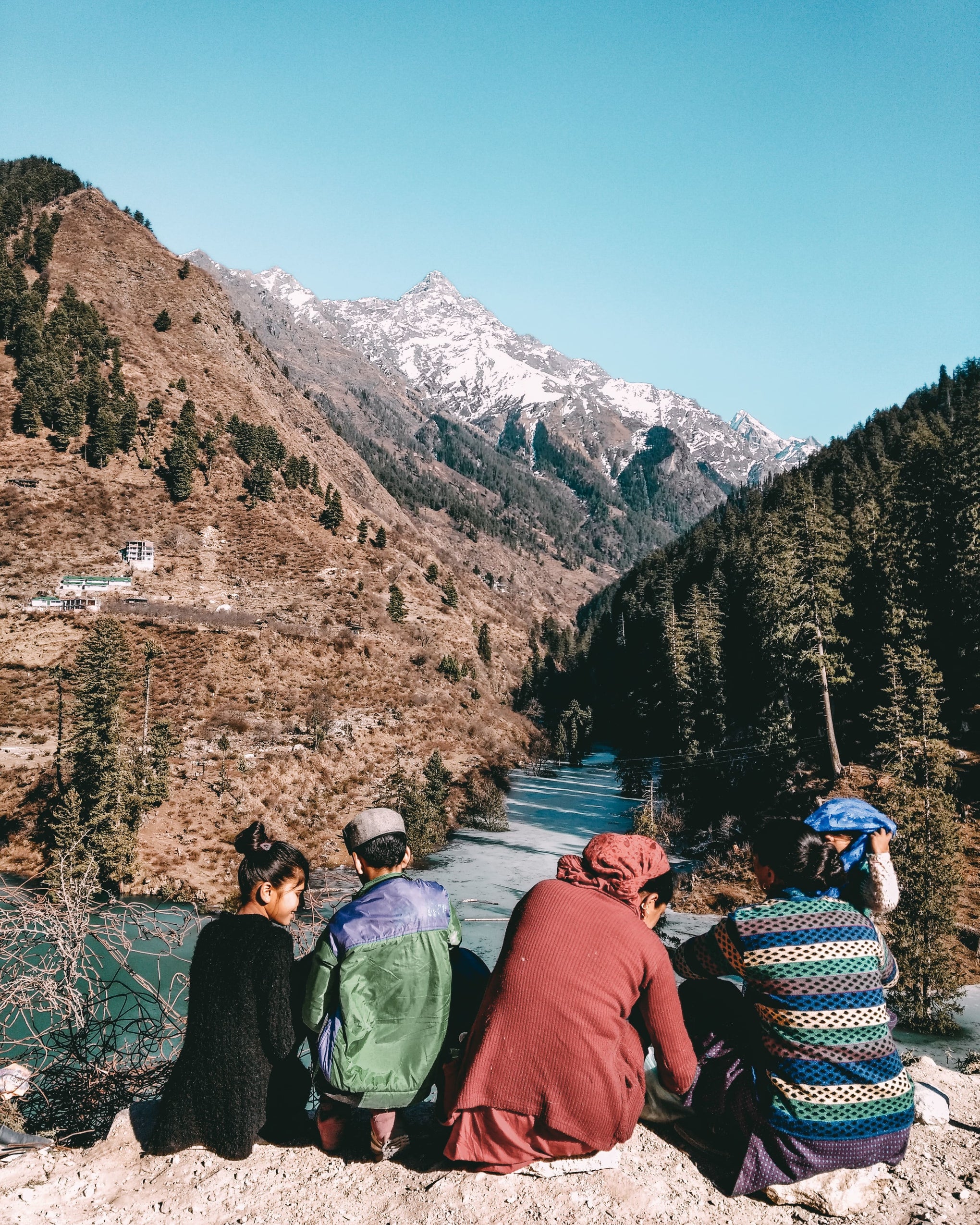 A group of younger folks sitting together looking over a mountain valley with colorful clothes on and a bit of snow in the peak of a mountain very far off in the distance. 