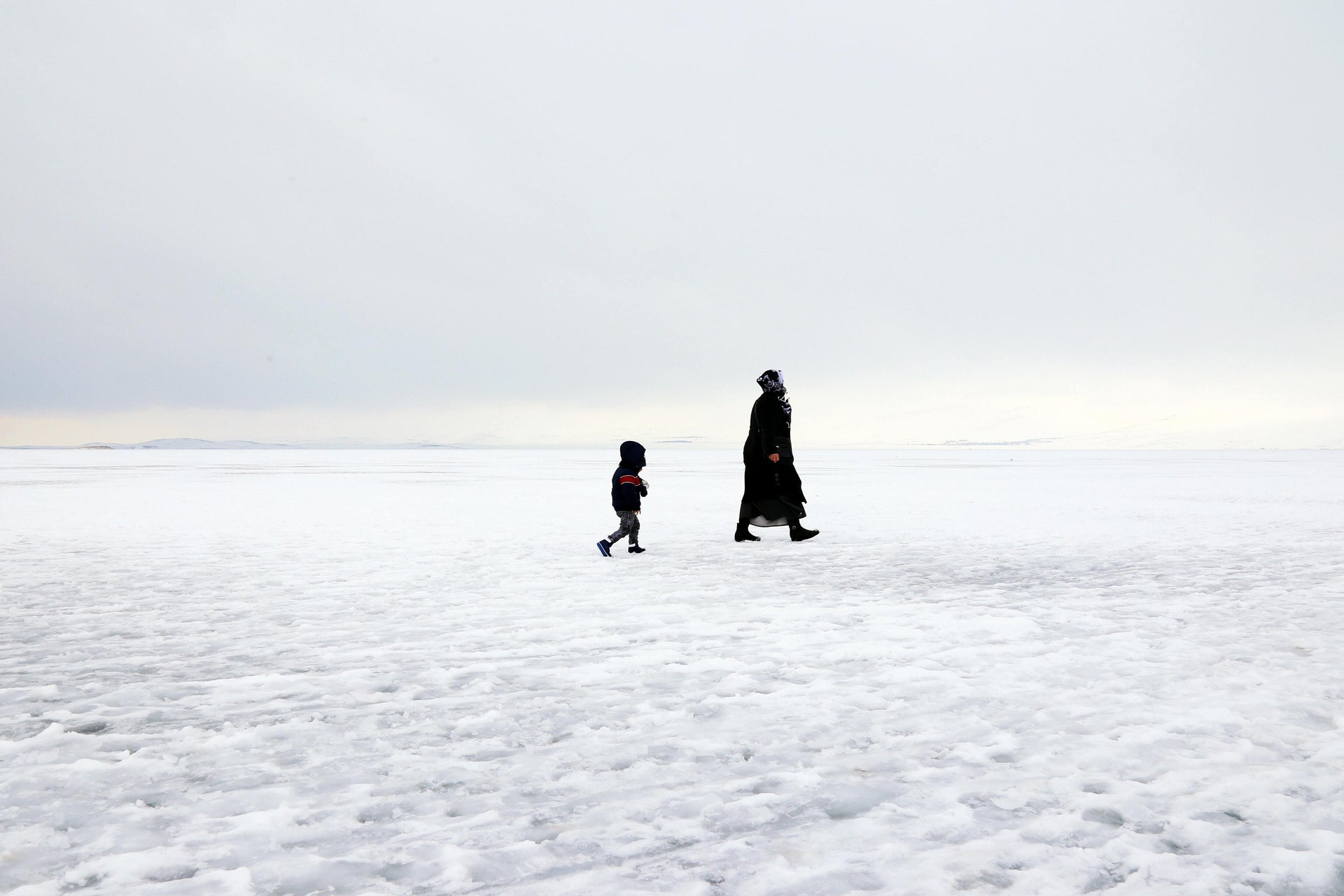 A Woman and a little boy in a pure white background with snow everywhere like a big plain of snow. They're walking alongside the perspective of the viewer as they brave the storm.