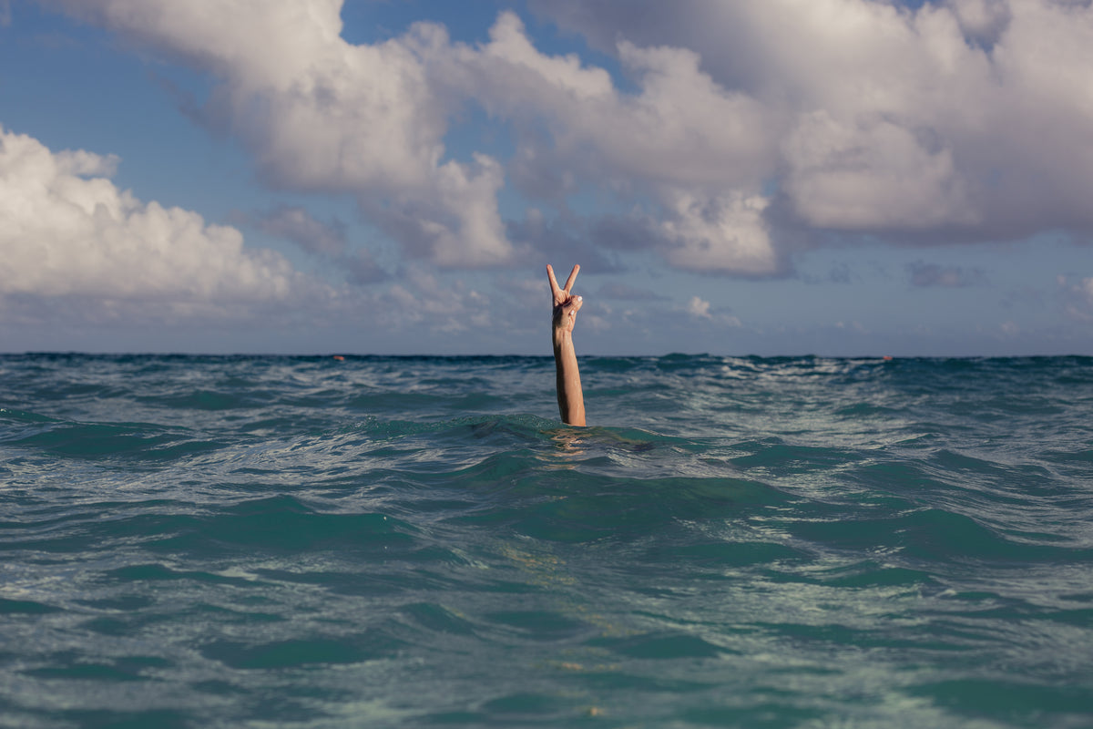 A girl with her hand sticking up at the sky in the blue in the deep ocean with clouds overhead and turquoise water consuming her, she still sticks her hand out of the water with a peace sign.