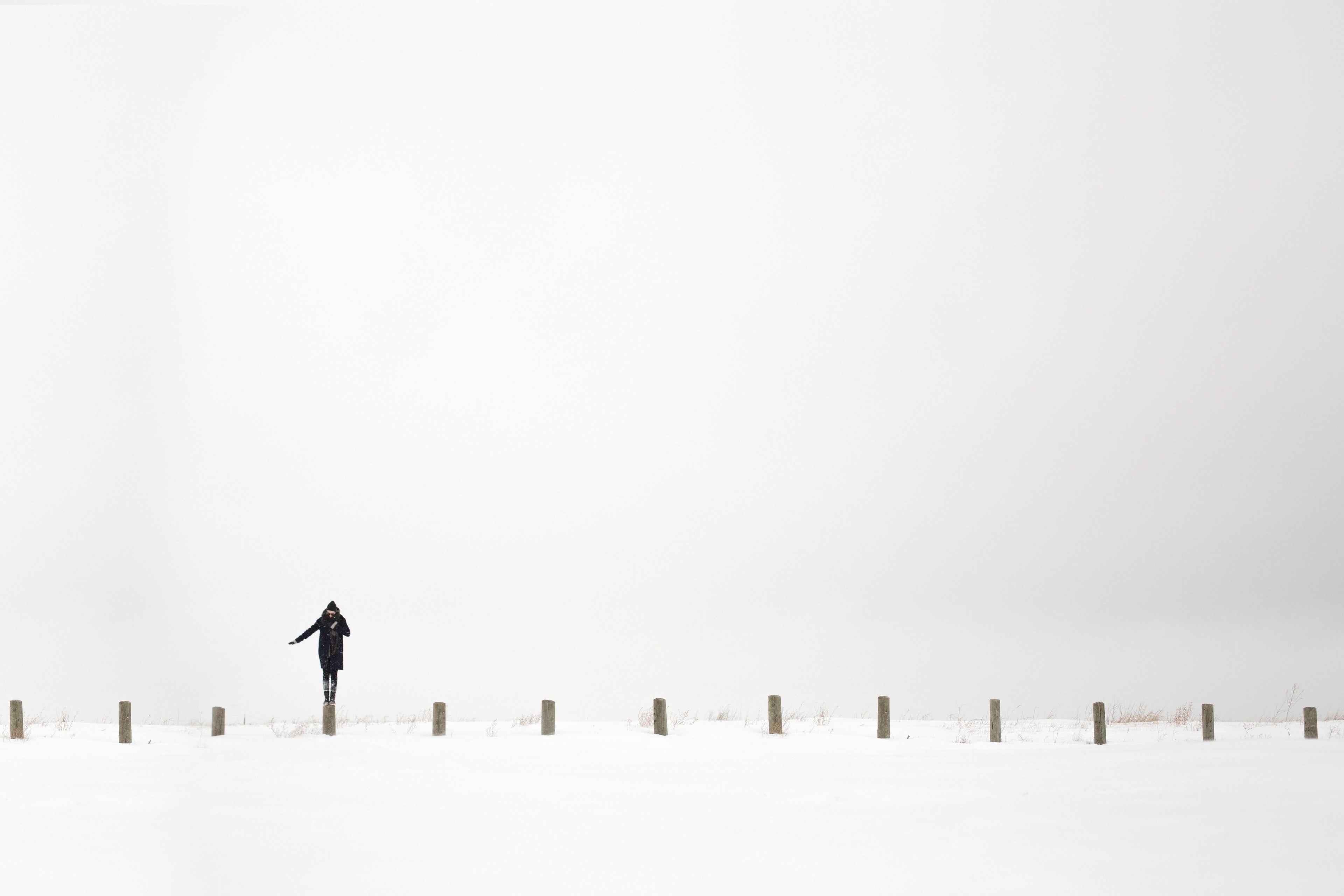 A woman Standing on one pole with her hand thrusted out at her side with nothing but blank snow around her everywhere and aside from that there are more of those wooden stakes like the one shes standing on.